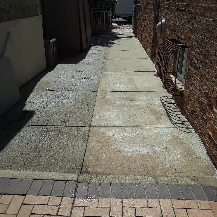 A concrete walkway between brick buildings displays a stark before-and-after contrast from pressure washing, with half the surface appearing clean and bright while the other half remains dark and stained.