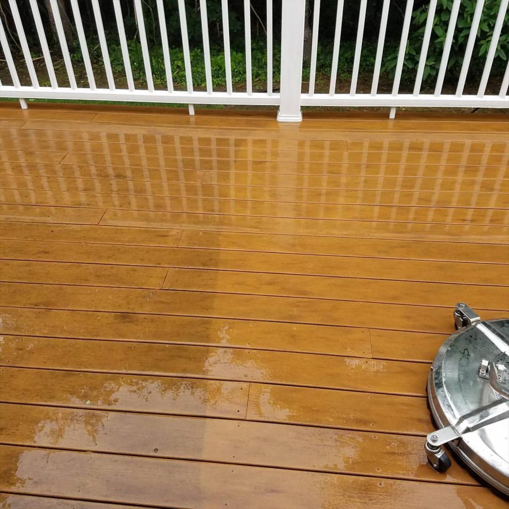 A wooden deck shows the dramatic results of pressure washing, with the upper portion appearing clean and golden while the lower section remains weathered and stained, alongside a visible pressure washing equipment.