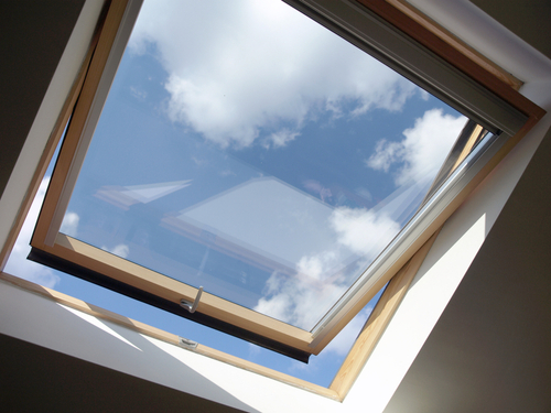 A wooden-framed skylight window is open, revealing a view of blue sky with white clouds from inside a room.