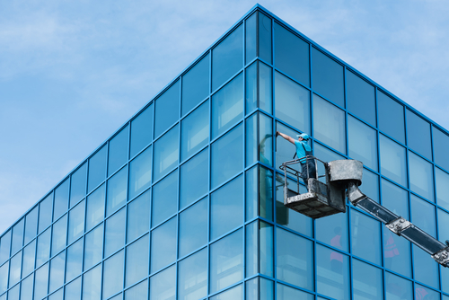 A worker in a mechanical lift platform is cleaning the exterior glass windows of a modern blue-tinted office building against a cloudy sky.RetryClaude can make mistakes. Please double-check responses.